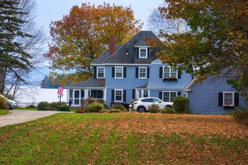 Blue colonial house with fall leaves on the lawn and a car in the driveway.
