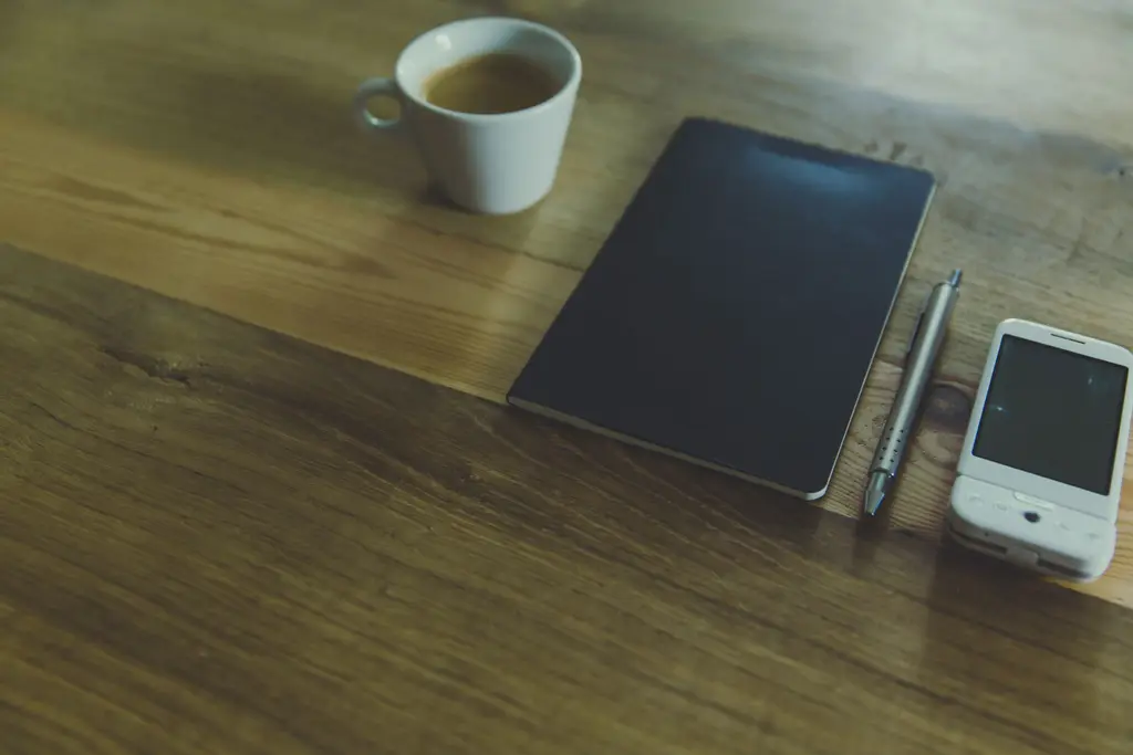 Phone, notebook, pen and a cup of coffee on a wooden table.