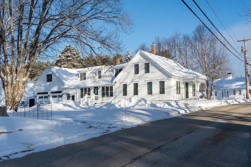 White clapboard New England farmhouse behind fresh snowbanks on a bright winter morning.