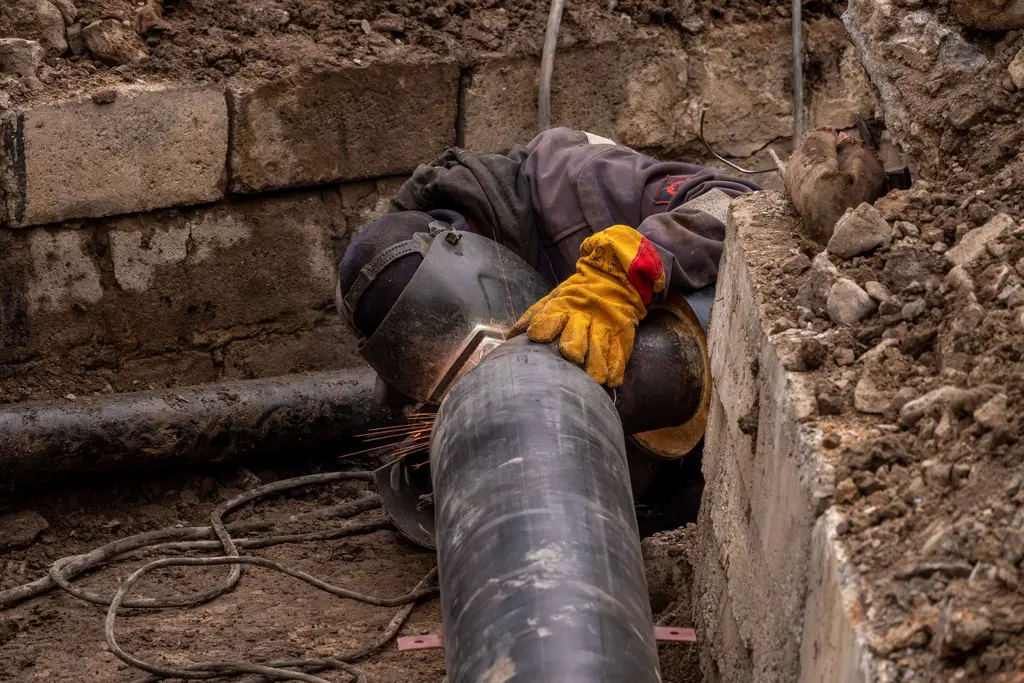 Worker in gloves on a pipe in a trench beside an old stone foundation.