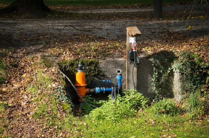 Open access pit in a yard with exposed pipe fittings and fallen leaves.